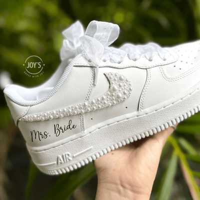 Custom white wedding Nike Air Force 1 with pearl-embellished swoosh, white ribbon laces, and "Mrs. Bride” on the side shown held in hand