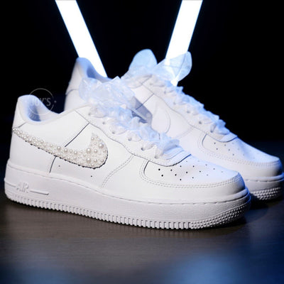 Custom white wedding Nike Air Force 1 with pearl-embellished swoosh, white ribbon laces, and personalized initials “J & E” on the heel, shown held in hand with soft bokeh lights in the background.